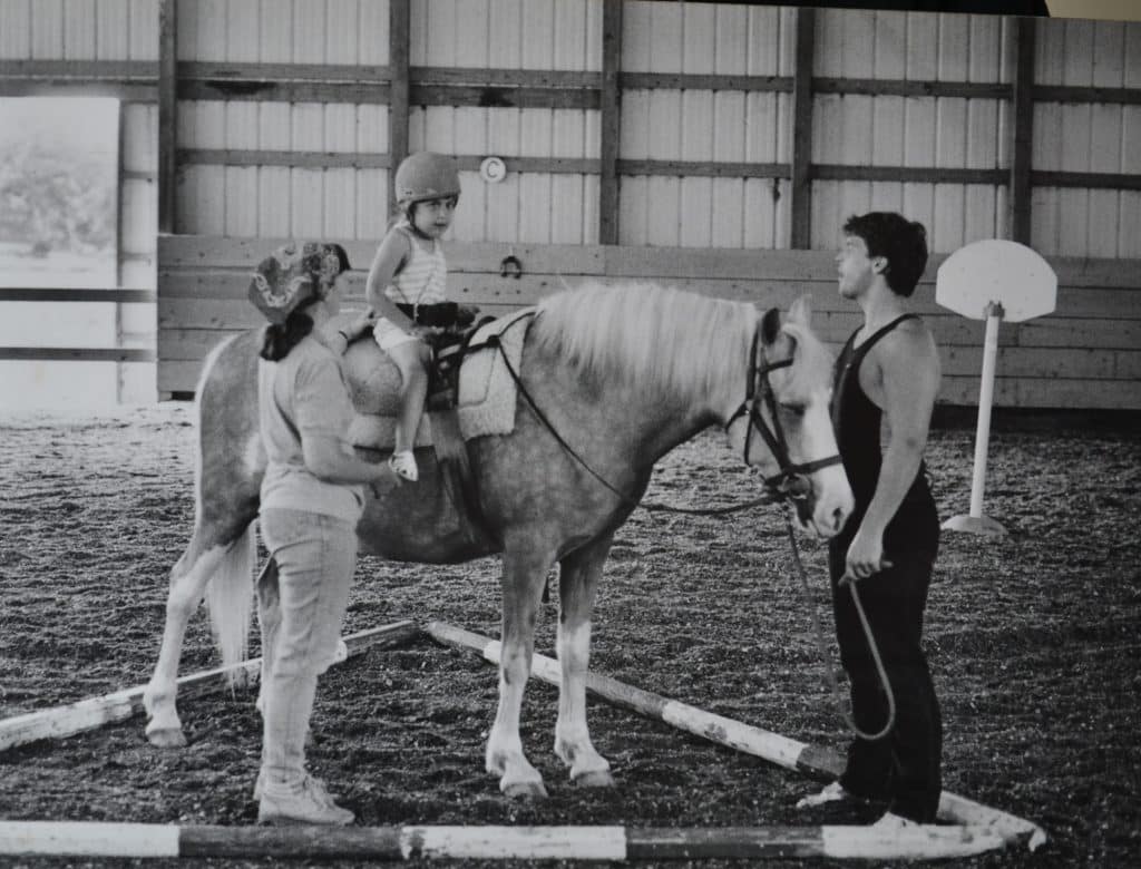 A young Sara Foszcz with a rider and side walker during an adaptive riding lesson.