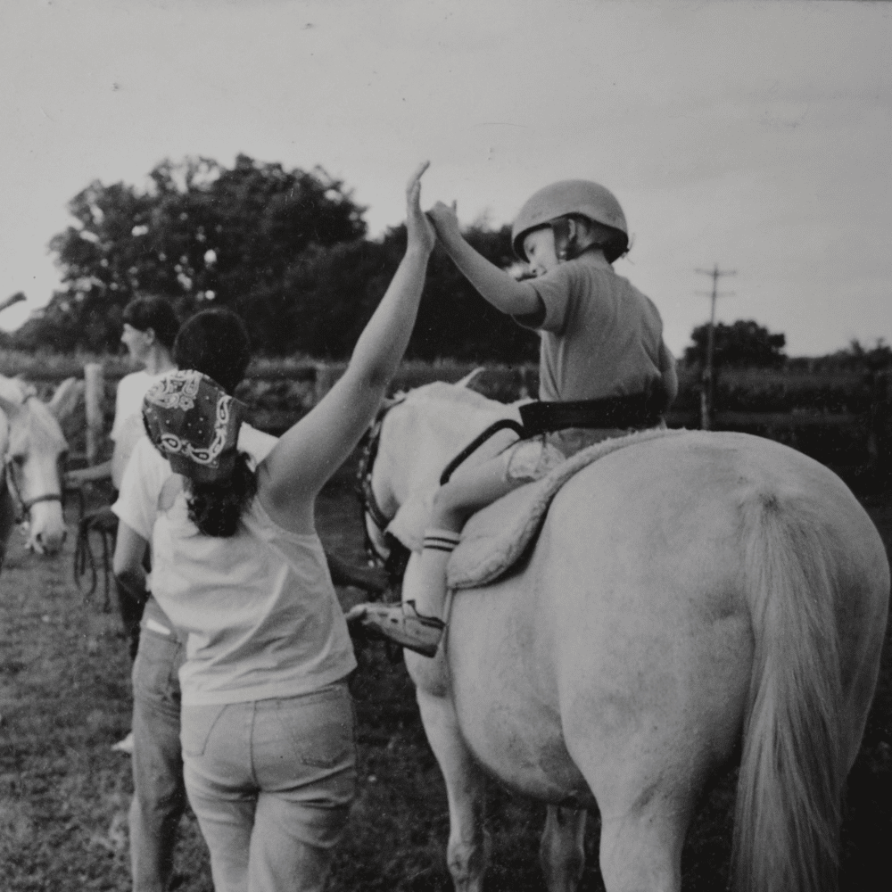 A young Sara Foszcz high-fives a rider during an adaptive riding lesson..