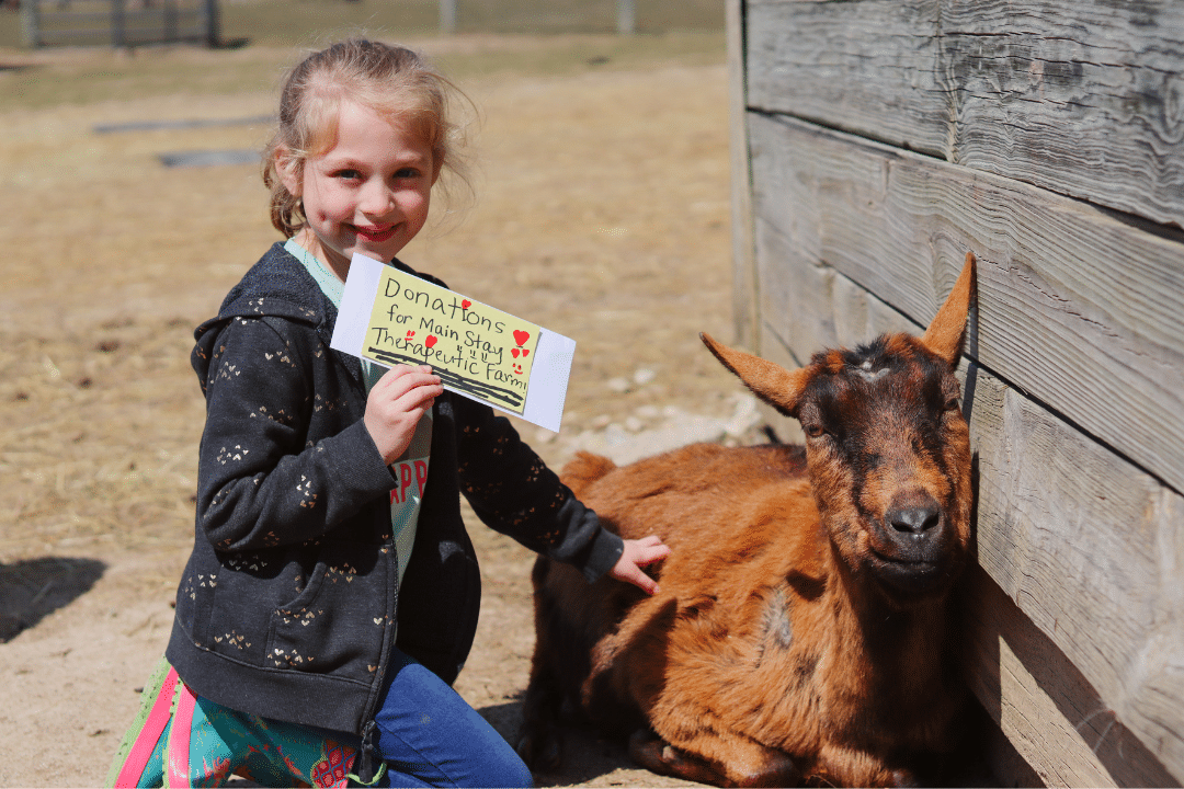 A young girl smiles while holding an envelope that says "Donations for Main Stay Therapeutic Farm" while smiling next to therapy goat Jasmin.