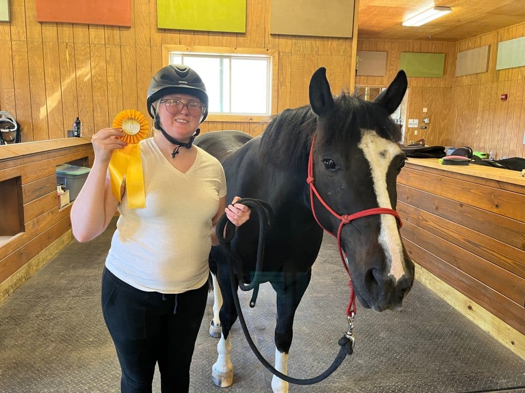 Rider Julia proudly holding up her Riders' Challenge ribbon while smiling next to her adaptive riding horse. 