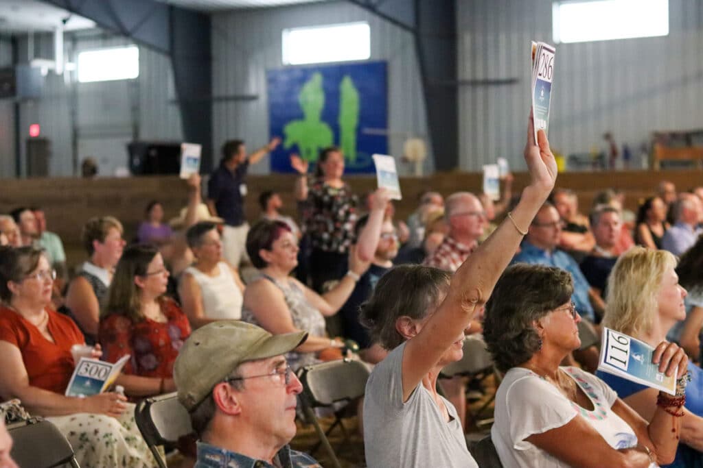 Audience members seated in an adaptive riding arena raise numbered paddles during a reverse auction, with rows of people watching and participating.